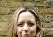 A picture of psychotherapist Lucy Lindop, who has long light brown hair and wears a black top, standing in front of a brick wall and looking directly at the camera with a neutral expression.