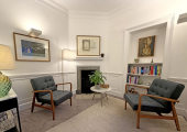 A calm, private psychotherapy room in the heart of Soho, thoughtfully designed for weekly sessions. The space features two mid-century armchairs facing each other, a small round table with a plant, artwork on the walls, a fireplace, and a bookshelf, 