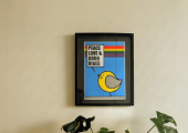 Monstera plant in a wicker plant pot holder, sitting on a white cabinet,next to a blue marbled decorative lamp, Above is a bold pop art style limited edition art print picture of a bird that is yellow, saying peace love and good vibes with a rainbow 
