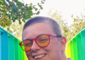 A person in glasses with very short hair is smiling. There is a rainbow painted bridge behind.