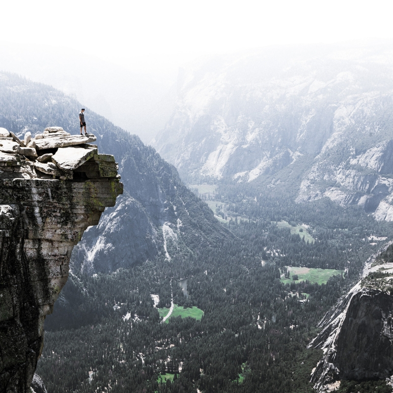 A person stands on top of a cliff edge, overlooking the valley below