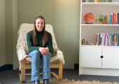 Bryony smiles from a chair in her counselling room, next to a bookcase.