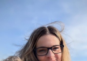 A smiling woman on the beach wearing glasses.