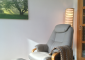This is an image of a comfortable, grey, recliner and footstool. The floor is wooden with a cream, shaggy rug. There is a large picture of green trees on the wall.