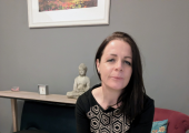 A lady say in a therapy room, on a chair and looking towards the camera.