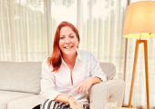 A woman with red hair sitting on asofa with her legs crossed wearing a white shirt and black and whi
