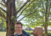 A young man smiling whilst standing up, whilst Simon works with him
